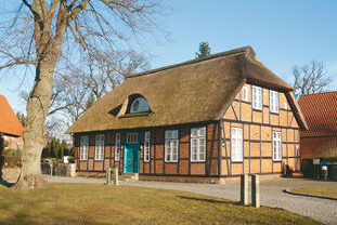 Historisches Küsterhaus mit Reetdach und Fachwerkfassade in Braun und Weiß, vor einem großen Baum.