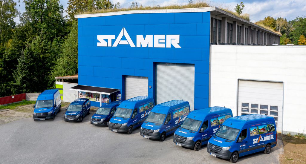Blaue Industriehalle der Firma Stamer mit großem weißen Logo und begrüntem Dach, davor eine Reihe blauer Firmenfahrzeuge.