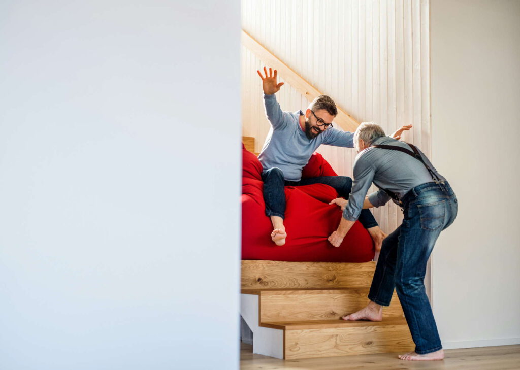 Zwei Männer interagieren spielerisch auf einer Holztreppe, wobei einer auf einem roten Sitzsack sitzt und der andere ihn festhält oder schiebt.