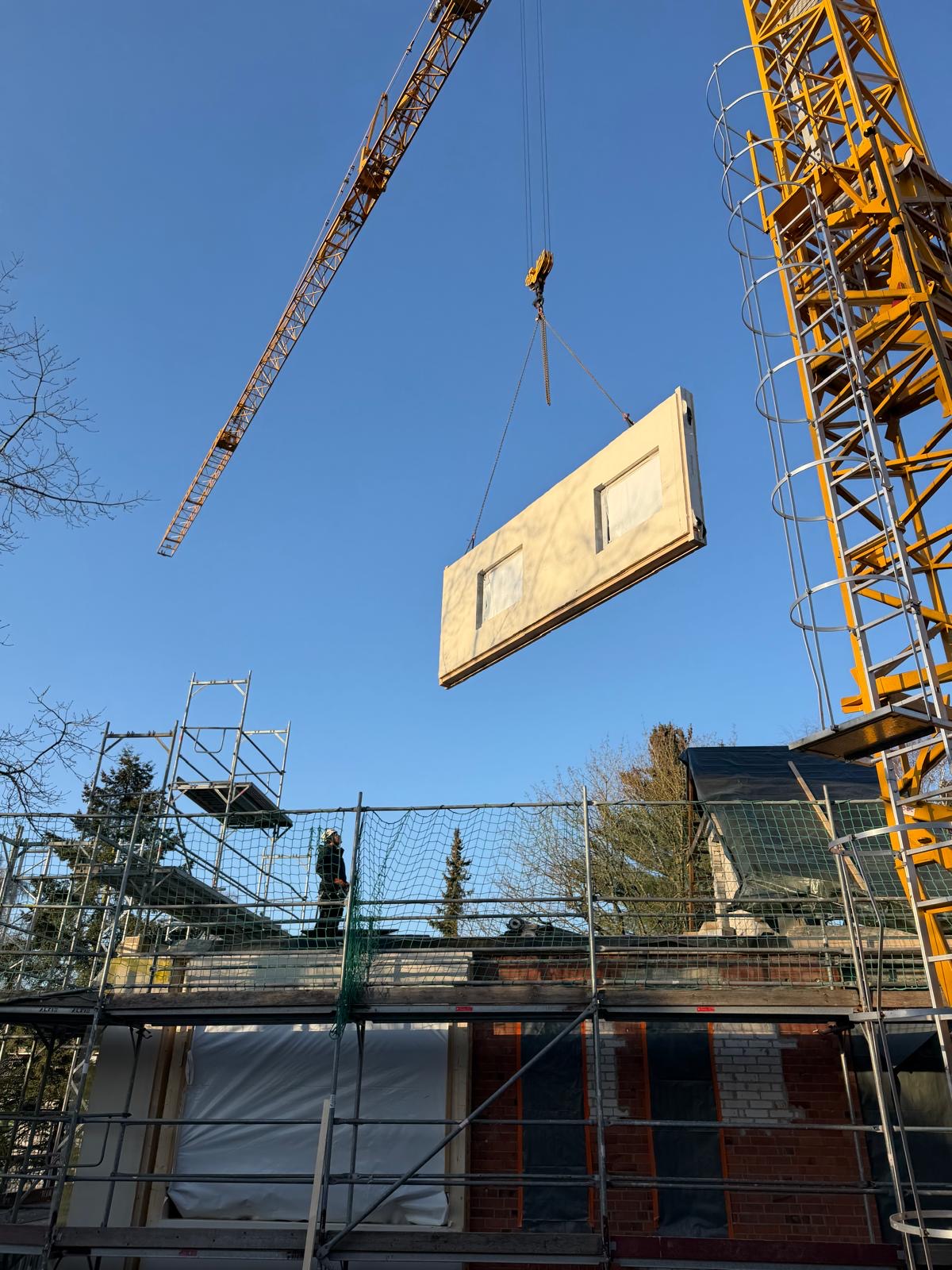 Kran hebt vorgefertigtes Fassadenelement über ein eingerüstetes Gebäude.