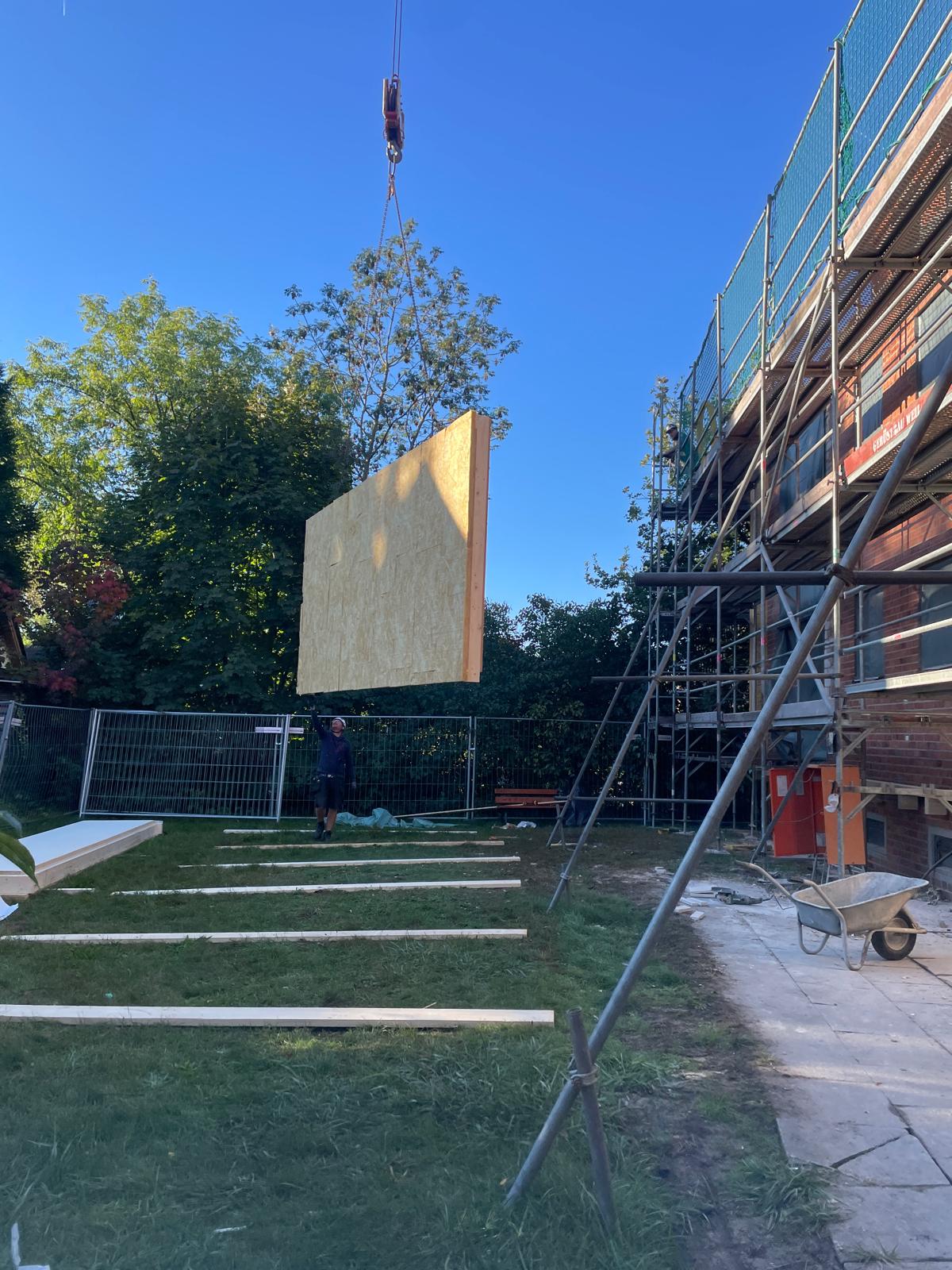 Kran hebt großes vorgefertigtes Wandelement auf einer Baustelle neben einem eingerüsteten Gebäude.