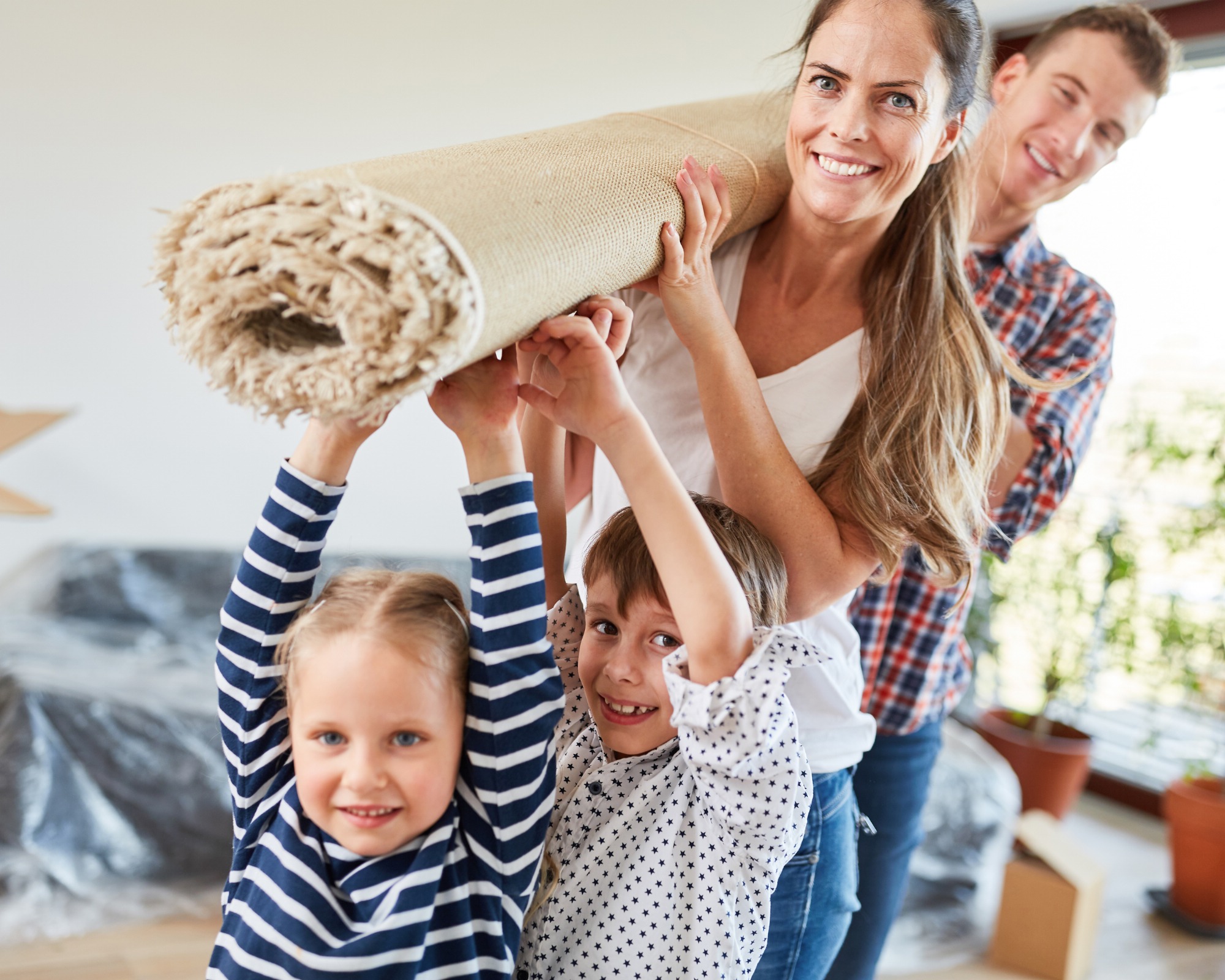 Familie mit zwei Kindern trägt gemeinsam einen zusammengerollten Teppich durch ein helles Zimmer, vermutlich beim Einrichten oder Umzug.