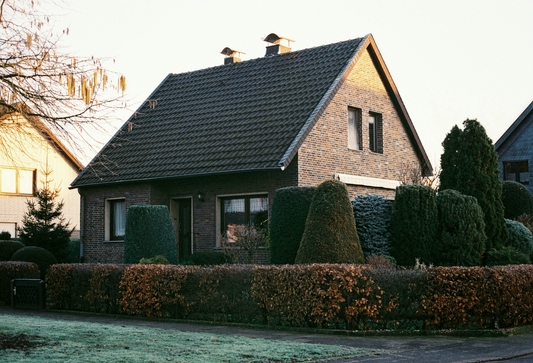 Einfamilienhaus mit Ziegeldach, Hecke und Vorgarten im Abendlicht.