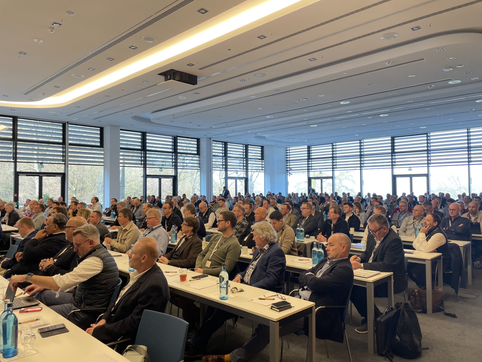 Großer Konferenzraum mit vielen sitzenden Teilnehmern, die aufmerksam einer Präsentation folgen; vor ihnen stehen Tische mit Notizen, Wasserflaschen und Arbeitsmaterialien.