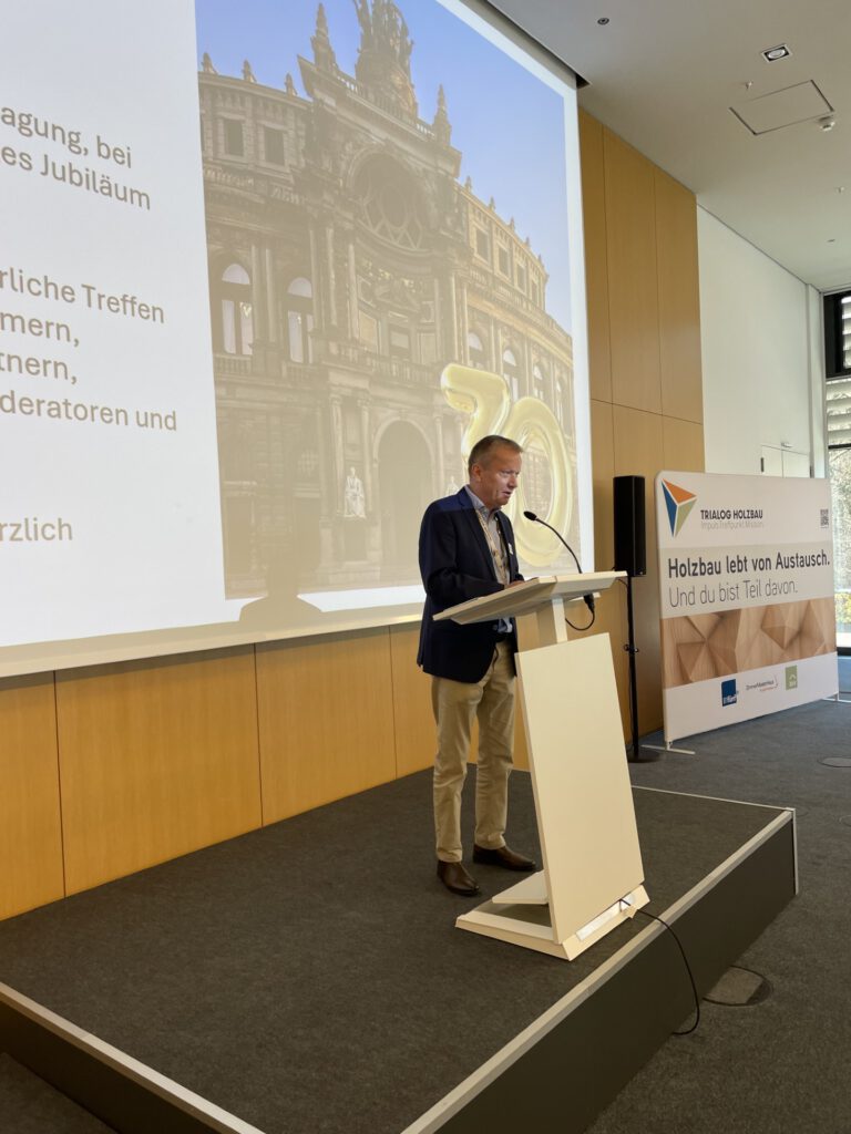 Ein Mann steht an einem Rednerpult auf einer Bühne und hält eine Präsentation; hinter ihm ist eine große Leinwand mit einer Gebäudedarstellung und Text, daneben ein Banner zum Thema Holzbau.